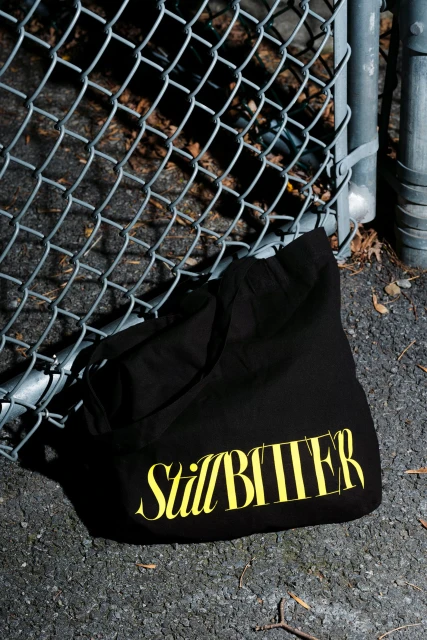 A black tote bag with yellow text rests against a chain-link fence on cracked asphalt outdoors, casting a shadow on a sunny day.