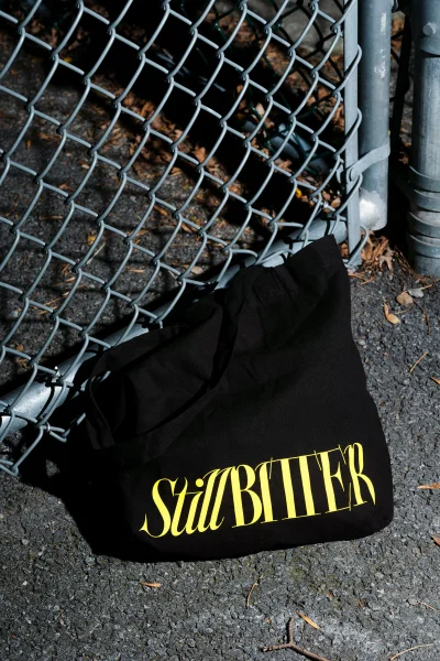 Black tote bag with yellow text resting on asphalt pavement in front of a metal chain-link fence