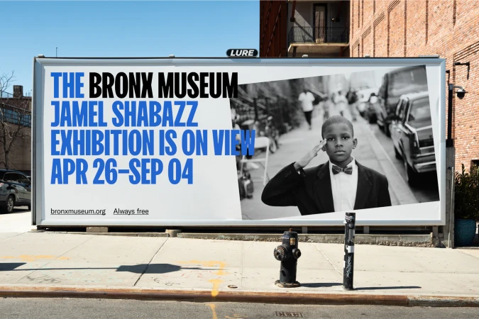Outdoor billboard featuring a black and white photo of a boy in a suit saluting with text promoting The Bronx Museum and Jamel Shabazz exhibition.