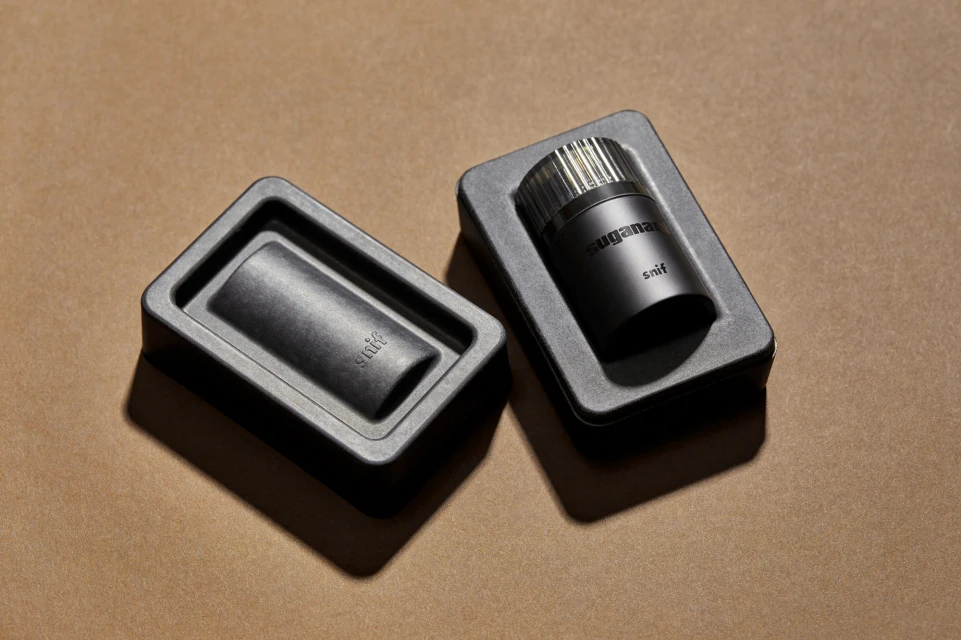 Two black boxes sit on a tan surface, one with a perfume bottle and one empty, showcasing product and packaging in a minimalist style.