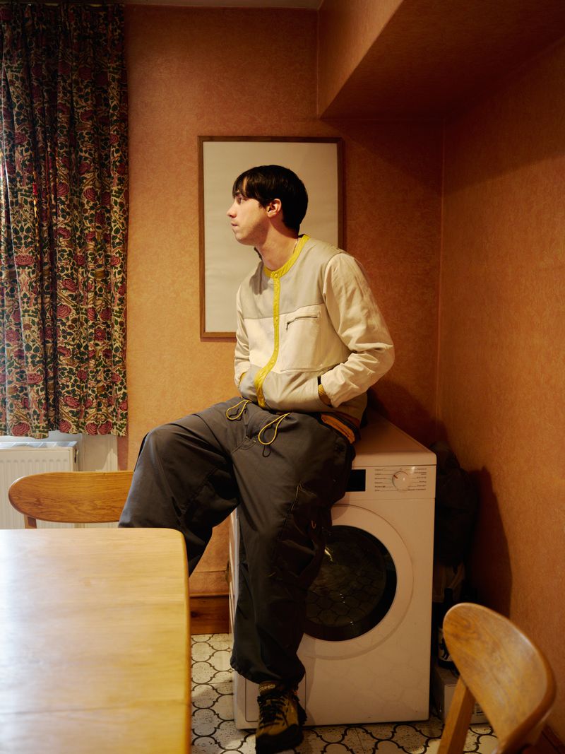 Young adult poses indoors wearing a jacket and casual pants, sitting on a washing machine in a peach-colored room with vintage patterned curtains.