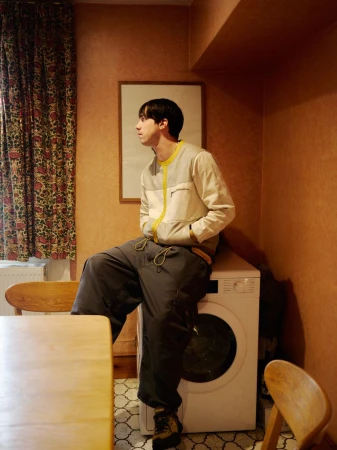 Young adult poses indoors wearing a jacket and casual pants, sitting on a washing machine in a peach-colored room with vintage patterned curtains.