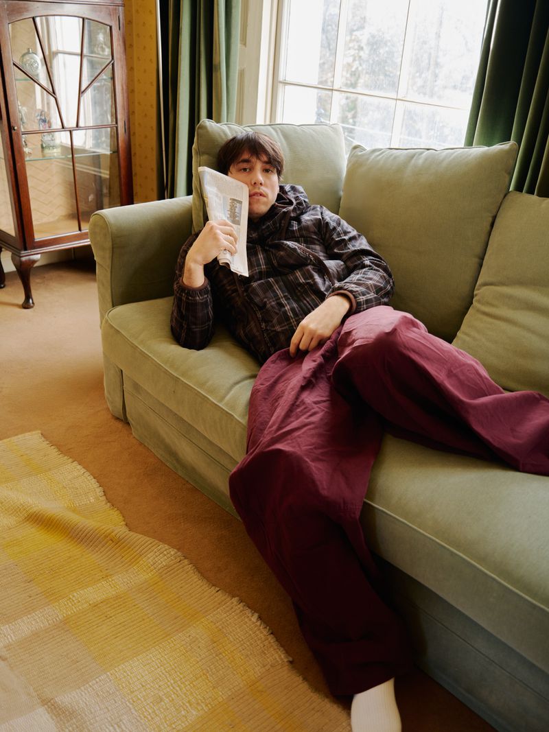 Man lounges on a couch, holding a newspaper in a living room with yellow patterned rug, wooden cabinet and window with green curtains.