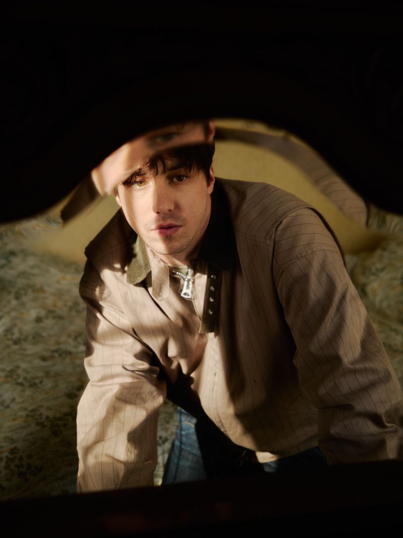 A medium shot captures an adult male looking up through a dark hole, wearing a collared tan shirt with blue jeans, indoors.