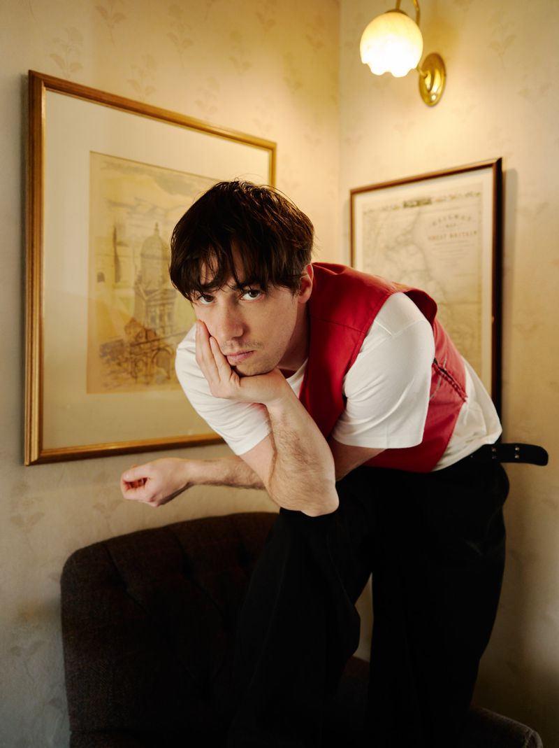 Portrait of a dark-haired man leaning on a chair in an indoor setting, wearing a white shirt and red vest, with framed pictures on the wall.