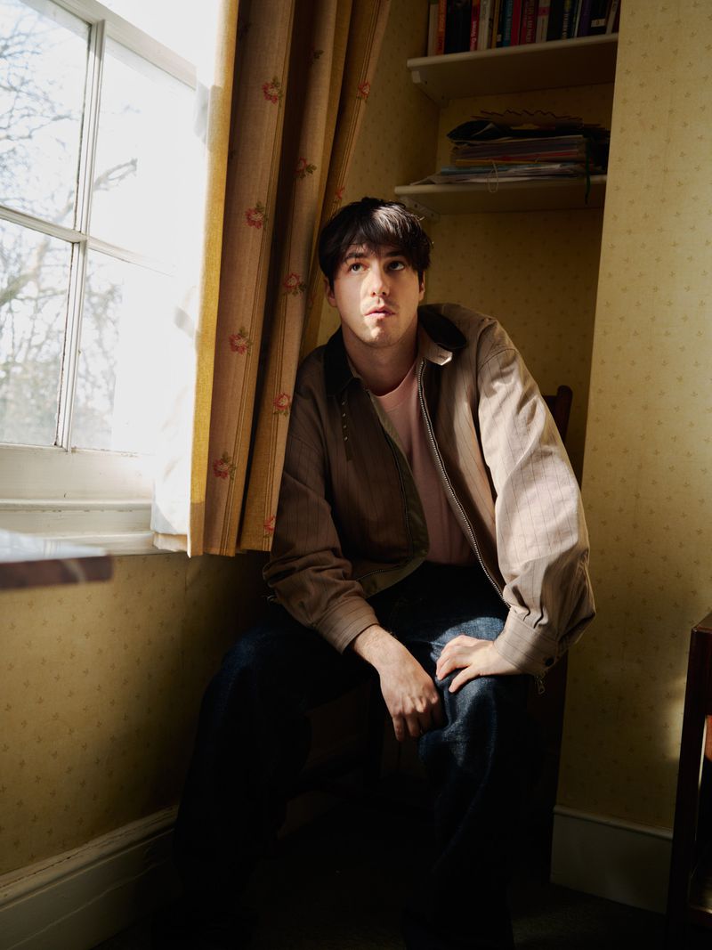 Man sits in sunlight by window in earth-toned jacket, pink shirt, dark jeans in room with floral wallpaper, shelves with books and magazines.