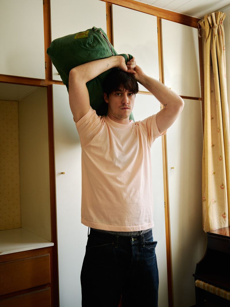 Young adult man carrying a large green bag over his shoulders indoors, wearing a pink t-shirt and dark jeans, with natural light.