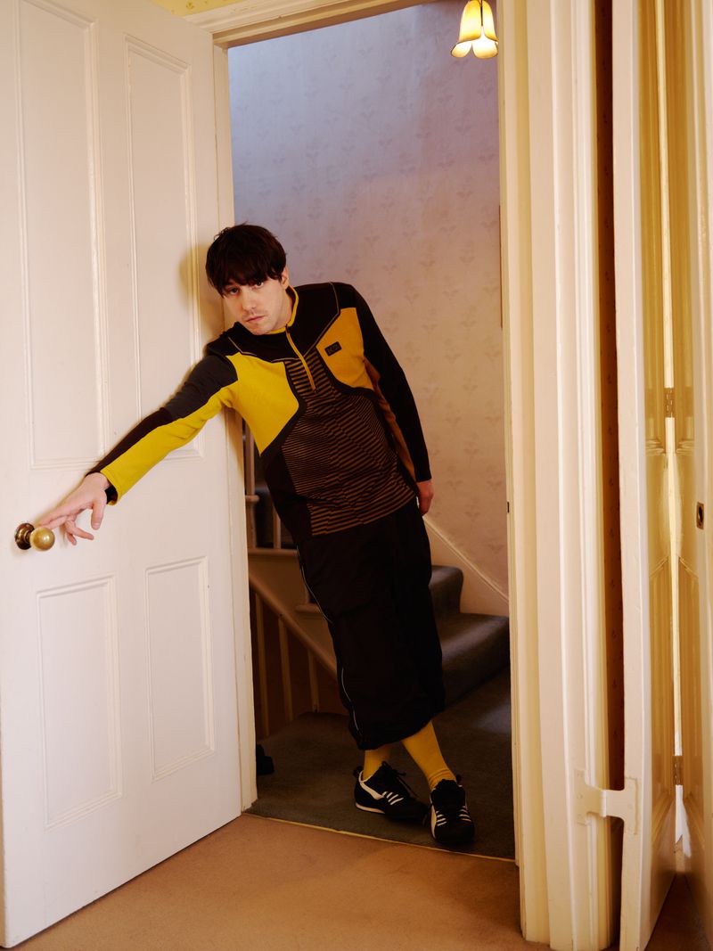 Indoor shot of a man in a black and yellow jacket, black skirt, and yellow socks leaning in a doorway with carpeted stairs visible.