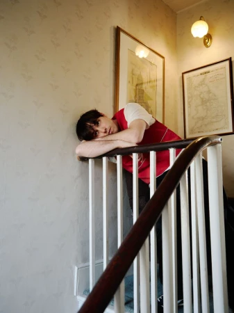 Portrait of a young adult male with dark hair leaning against a white staircase railing indoors, wearing a red and white vest.