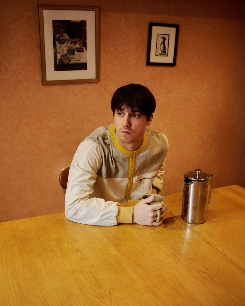 Man sitting at a wooden table, holding a mug, with framed art on the peach-colored wall, indoor lighting, medium shot, and neutral palette.