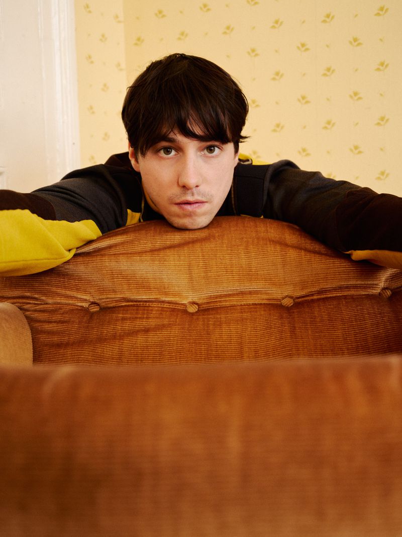 Portrait of a young man with dark hair leaning on the back of an upholstered armchair in an indoor setting with floral wallpaper.
