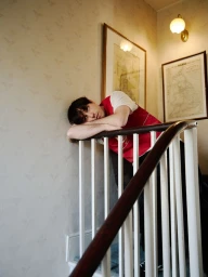 A young man leaning on a white wooden staircase railing in a patterned wallpaper hallway for Rollacoaster Magazine.