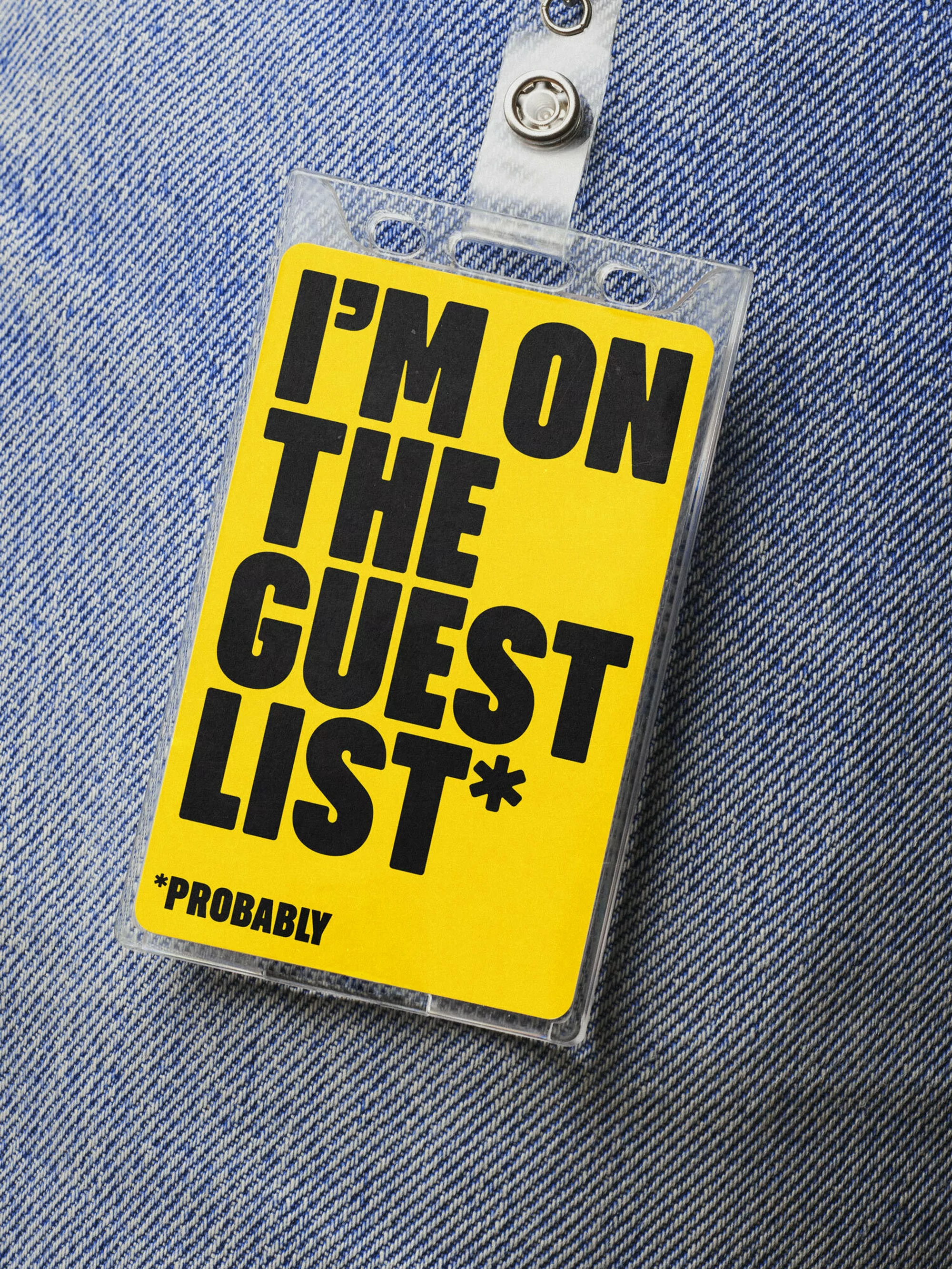 Close-up of a yellow guest pass with black sans-serif font reading 'I'm on the guest list* *probably' attached to denim clothing with silver snaps.