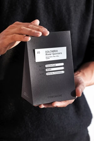 Hands holding a dark grey coffee bag with a label displaying 'Colombia Rivier Quintero,' emphasizing minimalist branding and product presentation.