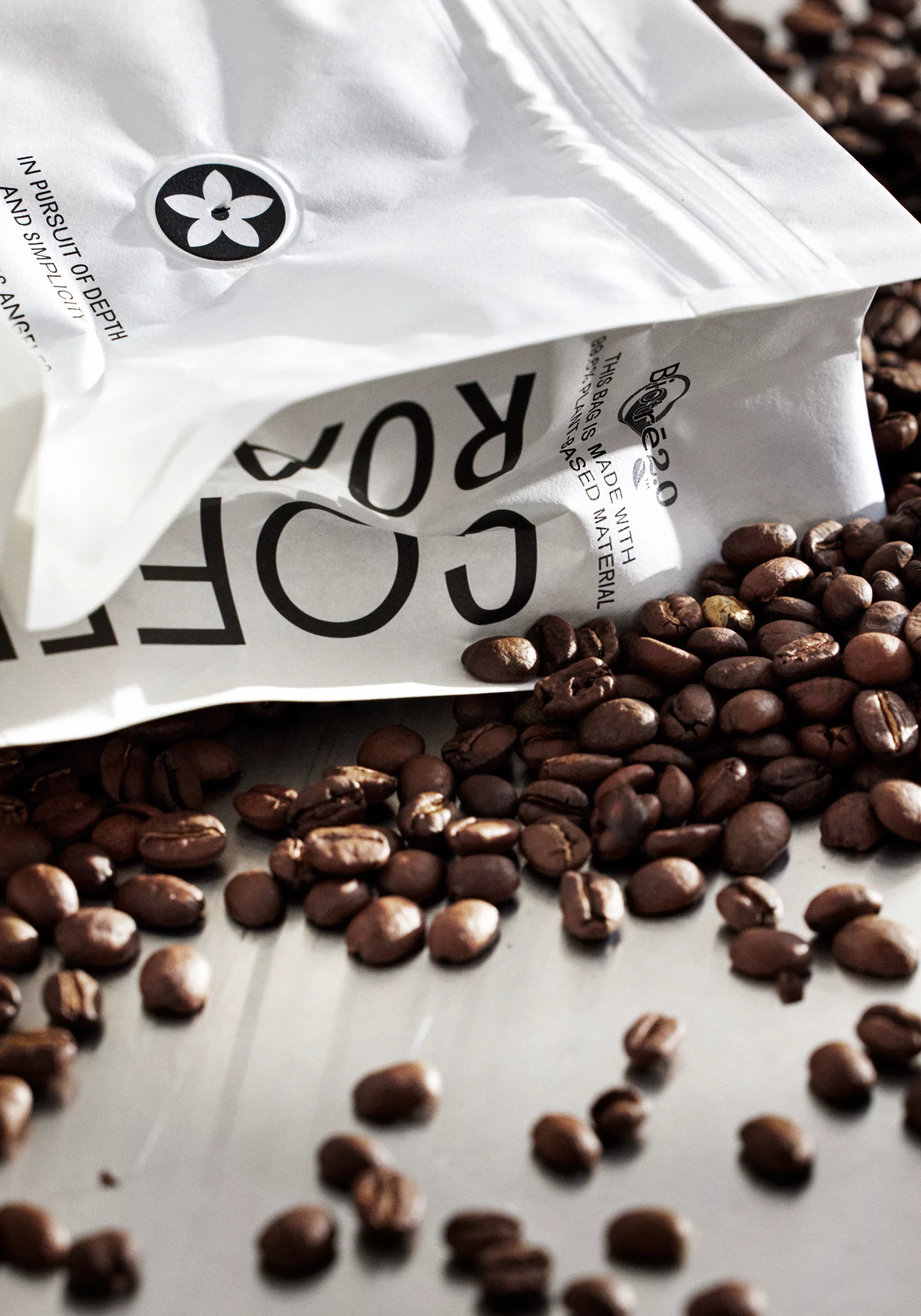 Close-up of roasted coffee beans scattered around a white coffee bag with black text on a reflective silver surface.