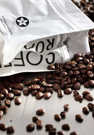 Close-up of roasted coffee beans scattered around a white coffee bag with black text on a reflective silver surface.