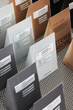 Close-up of triangle-shaped, grey, brown, and white coffee sample packages with labels on a metallic perforated surface.