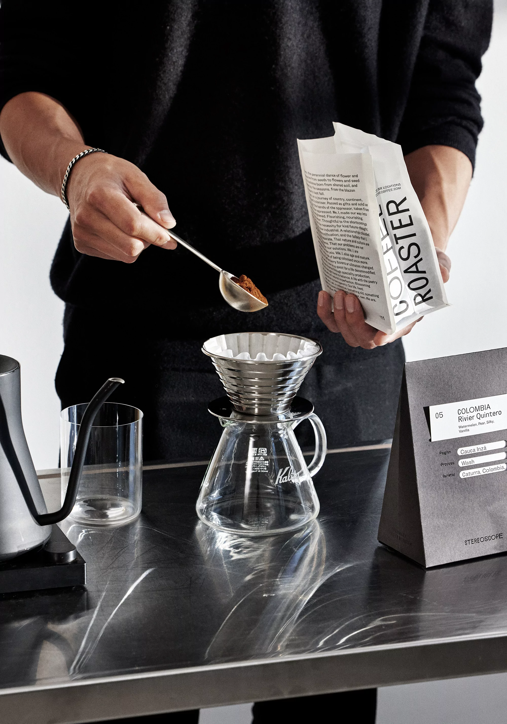 Coffee brewing process featuring a person using a spoon to scoop coffee grounds into a filter over a glass carafe on a metal table.