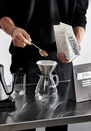 Person in a black sweater pouring coffee grounds into a glass dripper holding a coffee bag for Stereoscope brand identity