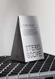 Pyramid-shaped grey paper packaging with embossed black typography resting on a dark plastic crate against a plain light wall