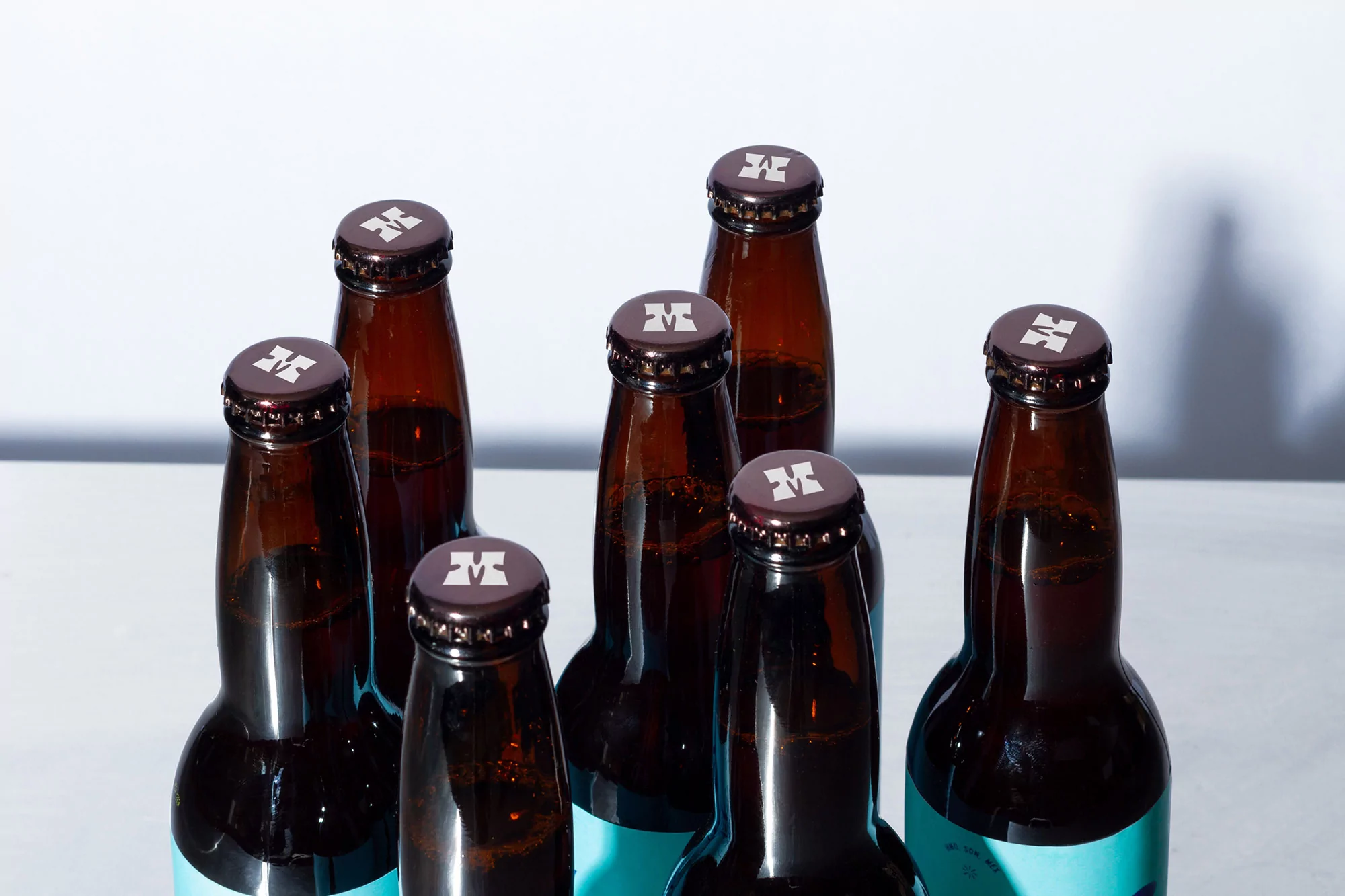Seven brown glass beer bottles with light blue labels and metal caps displaying a white logo on a white reflective surface.