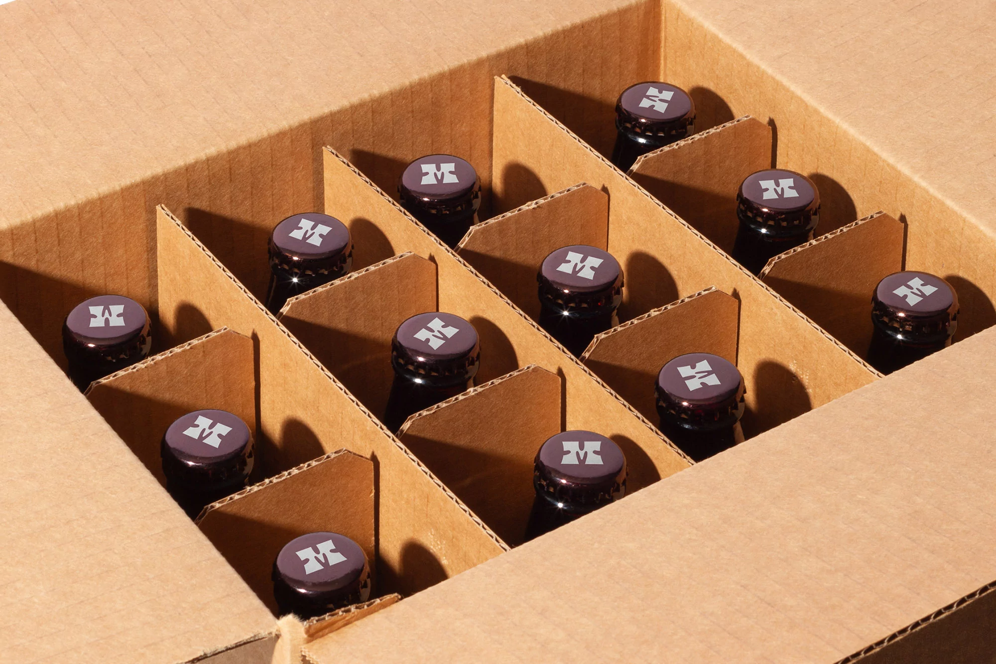 A box with cardboard dividers contains a neatly arranged collection of dark glass bottles capped with burgundy lids bearing a logo.