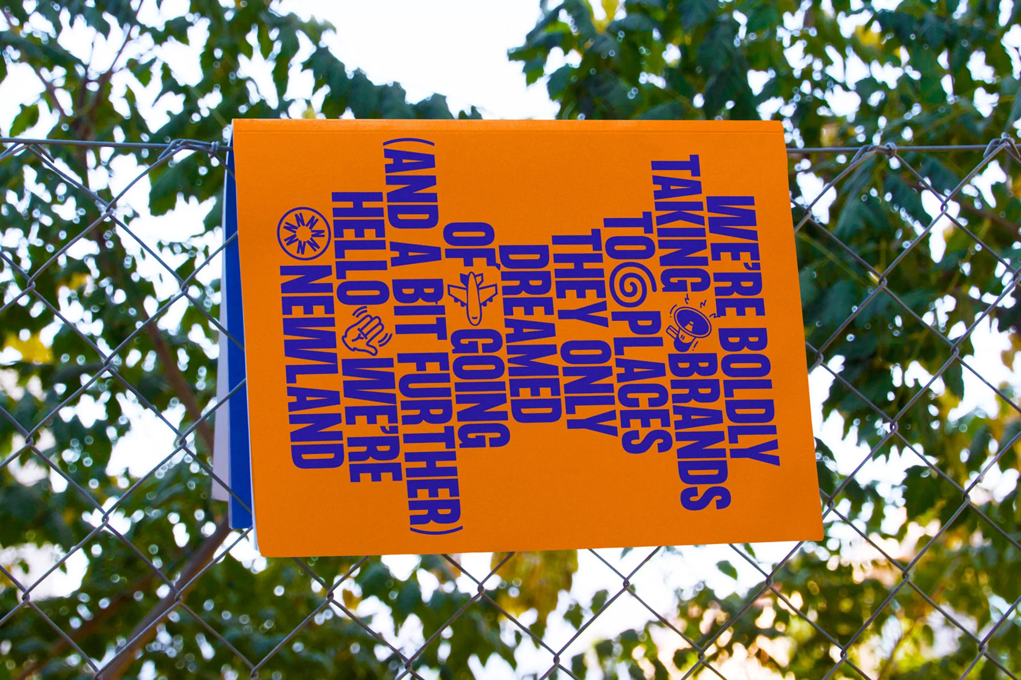 Orange sign on a chain-link fence with blue lettering that says, "We're boldly taking brands to places they only dreamed of going."