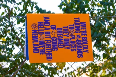 Orange sign with blue typography attached to a wire mesh fence against green foliage.