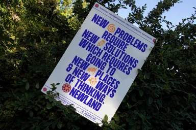White promotional poster with blue typography mounted amongst green leafy foliage outdoors.
