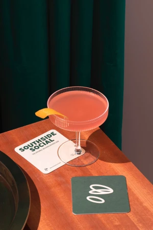 Pink cocktail in a coupe glass with a lemon twist on a wooden table next to a coaster and green curtains. 