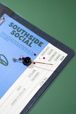 Cocktail cherry pierced by a metal needle on a blue menu with Southside Social branding against a green background