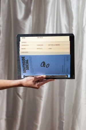 Hand holding a black binder with blue menu insert and tabbed divider cards against a draped white curtain background.