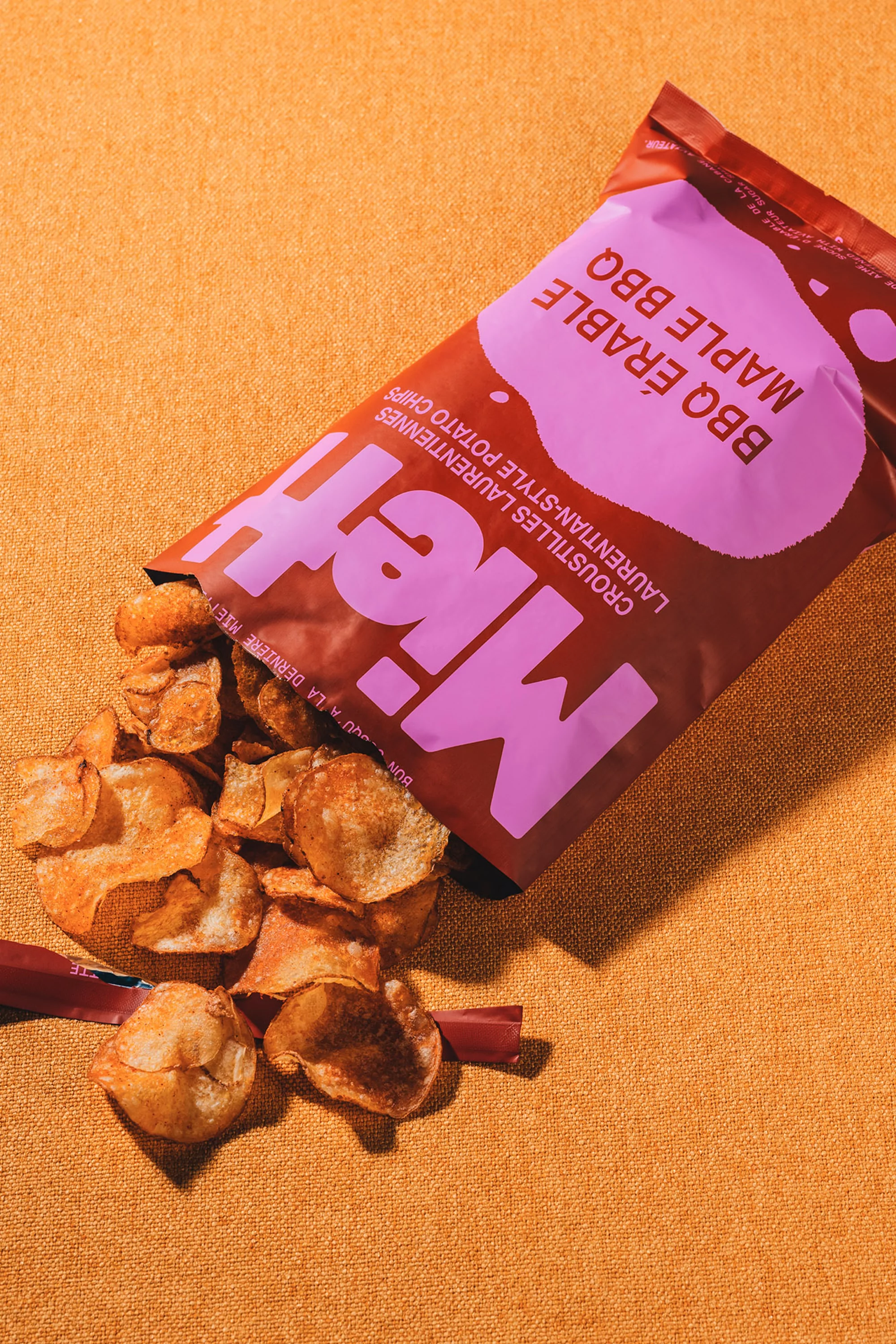 Bag of maple BBQ chips spilling onto a textured gold surface. Package is brown with a light purple logo.