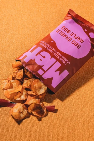 Bag of maple BBQ chips spilling onto a textured gold surface. Package is brown with a light purple logo.