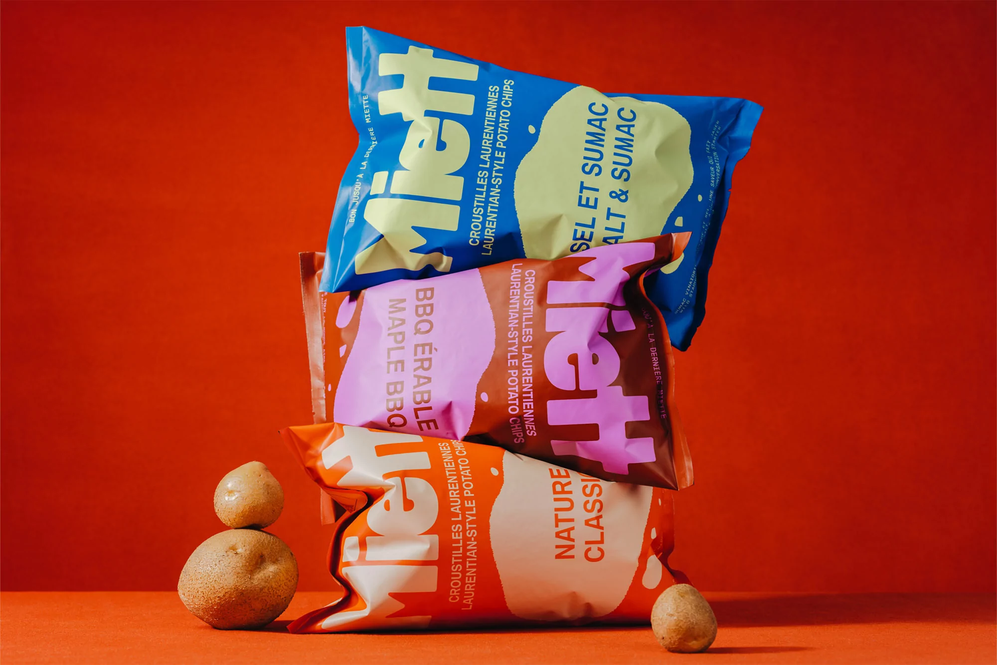 Three colorful bags of Miett Laurentian-Style Potato Chips stacked with two whole potatoes against an orange background.
