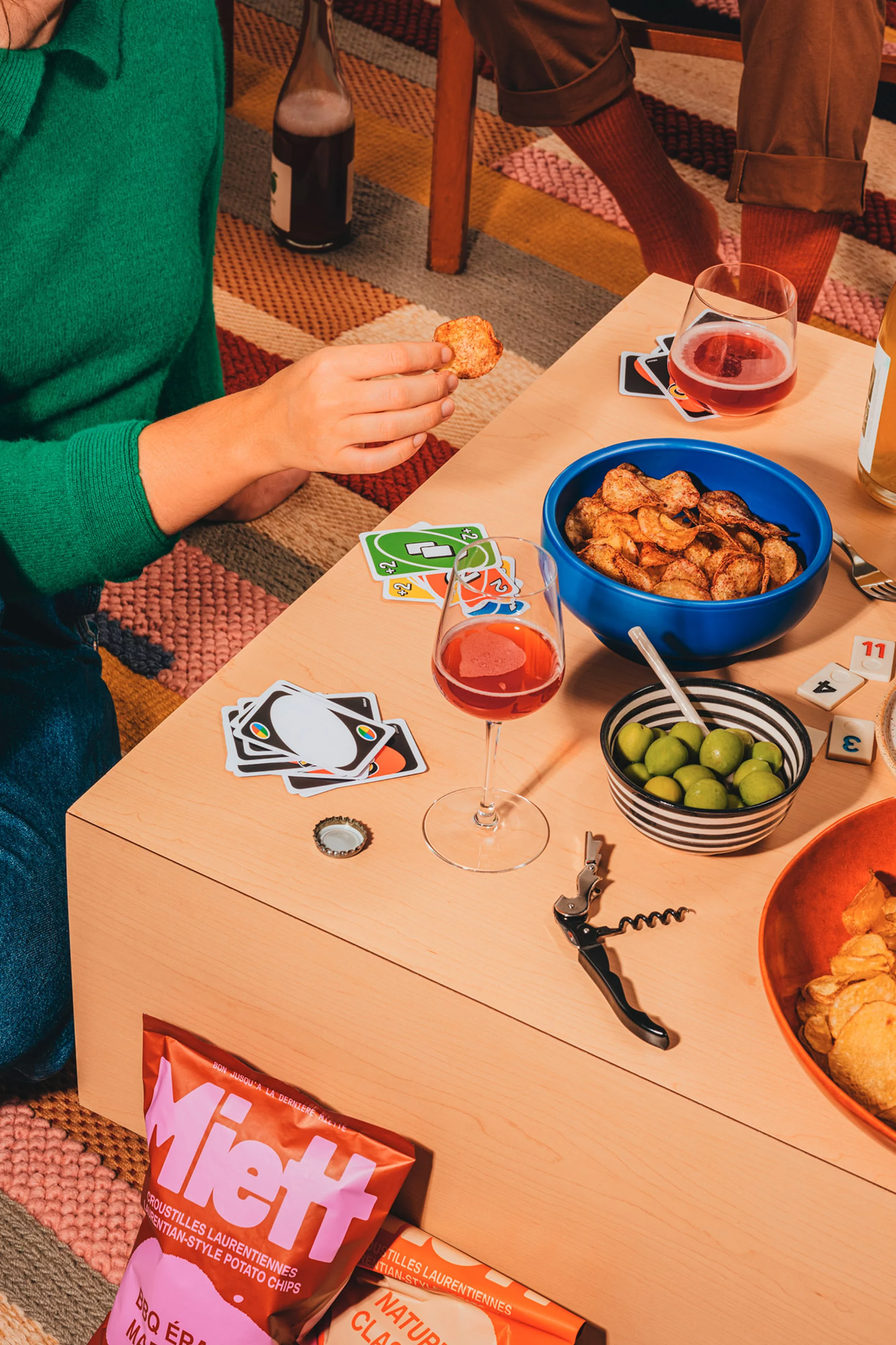Overhead shot of tabletop arrangement with snacks, drinks, playing cards, and two people seated around it on a patterned rug.