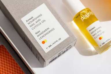 A bottle of nail and cuticle oil next to its cardboard packaging on a white and patterned orange surface.