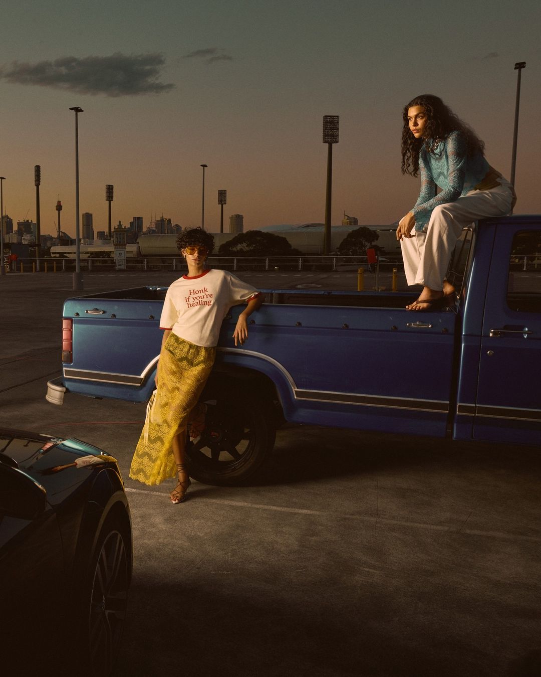 Two women pose near a blue pickup truck in an outdoor parking lot at dusk. One sits on the truck bed, while the other leans against it.