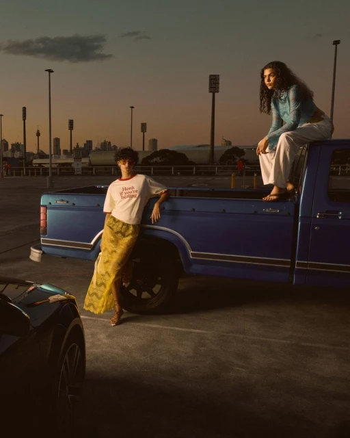Two women pose near a blue pickup truck in an outdoor parking lot at dusk. One sits on the truck bed, while the other leans against it.
