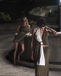Two women posing on a concrete structure at night, wearing olive green dresses with sunglasses.