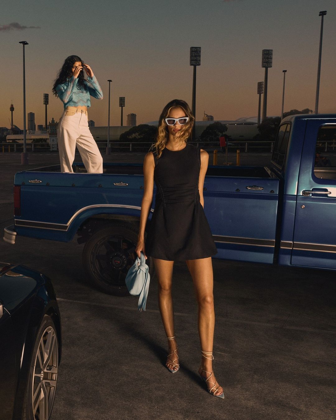 Two women pose in a parking lot near a blue pickup truck under a dusky sky. One woman is in the truck bed.
