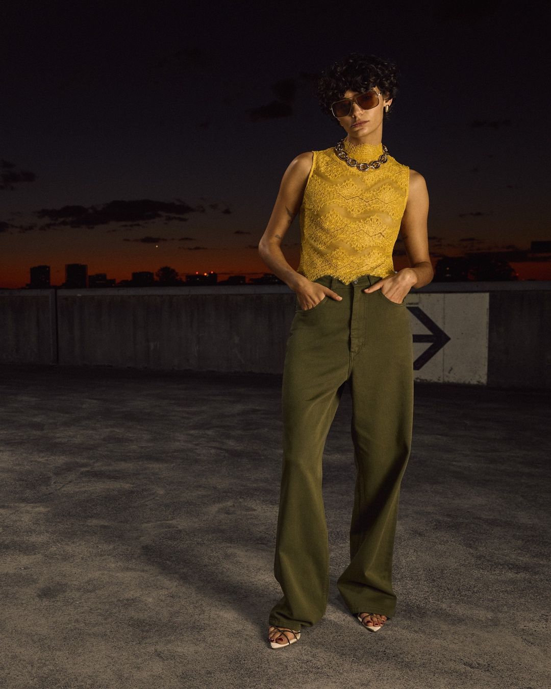 A young adult woman poses in yellow lace top and olive-green pants against an evening skyline and dark urban background.