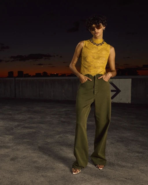 A young adult woman poses in yellow lace top and olive-green pants against an evening skyline and dark urban background.