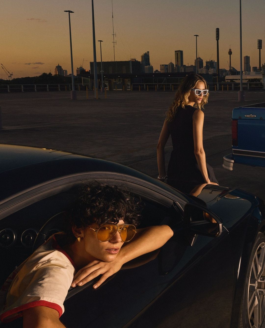 Two models outdoors at dusk with cars, one person leaning out of a car window wearing tinted sunglasses.