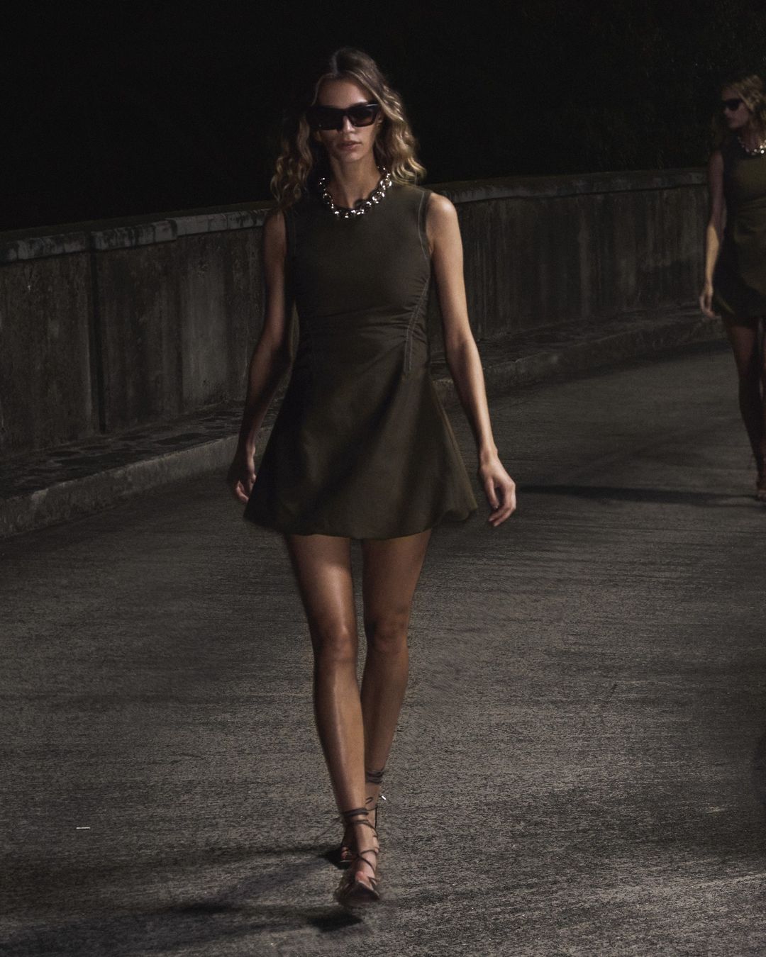 Two adult women walk on a street at night, showcasing dark minidresses, statement necklaces, and strappy heels in a dimly lit setting.