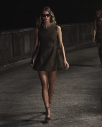 Model walking on an outdoor path wearing a dark sleeveless dress and sunglasses