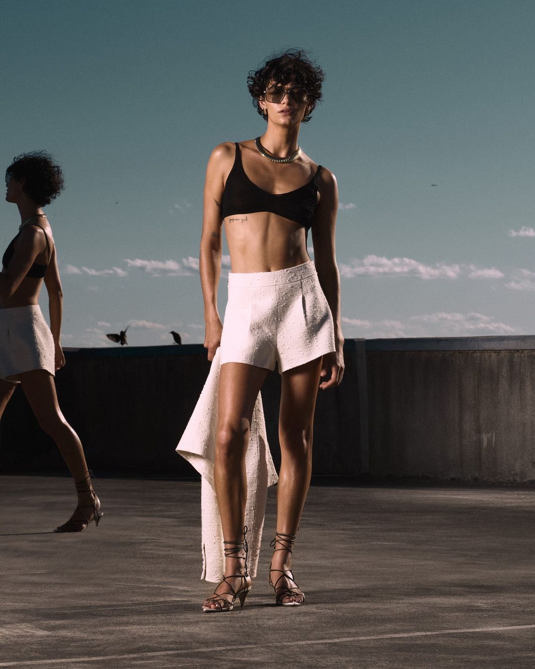 Two women on a rooftop wearing black tops and white shorts. One stands looking at the camera, while the other walks away.