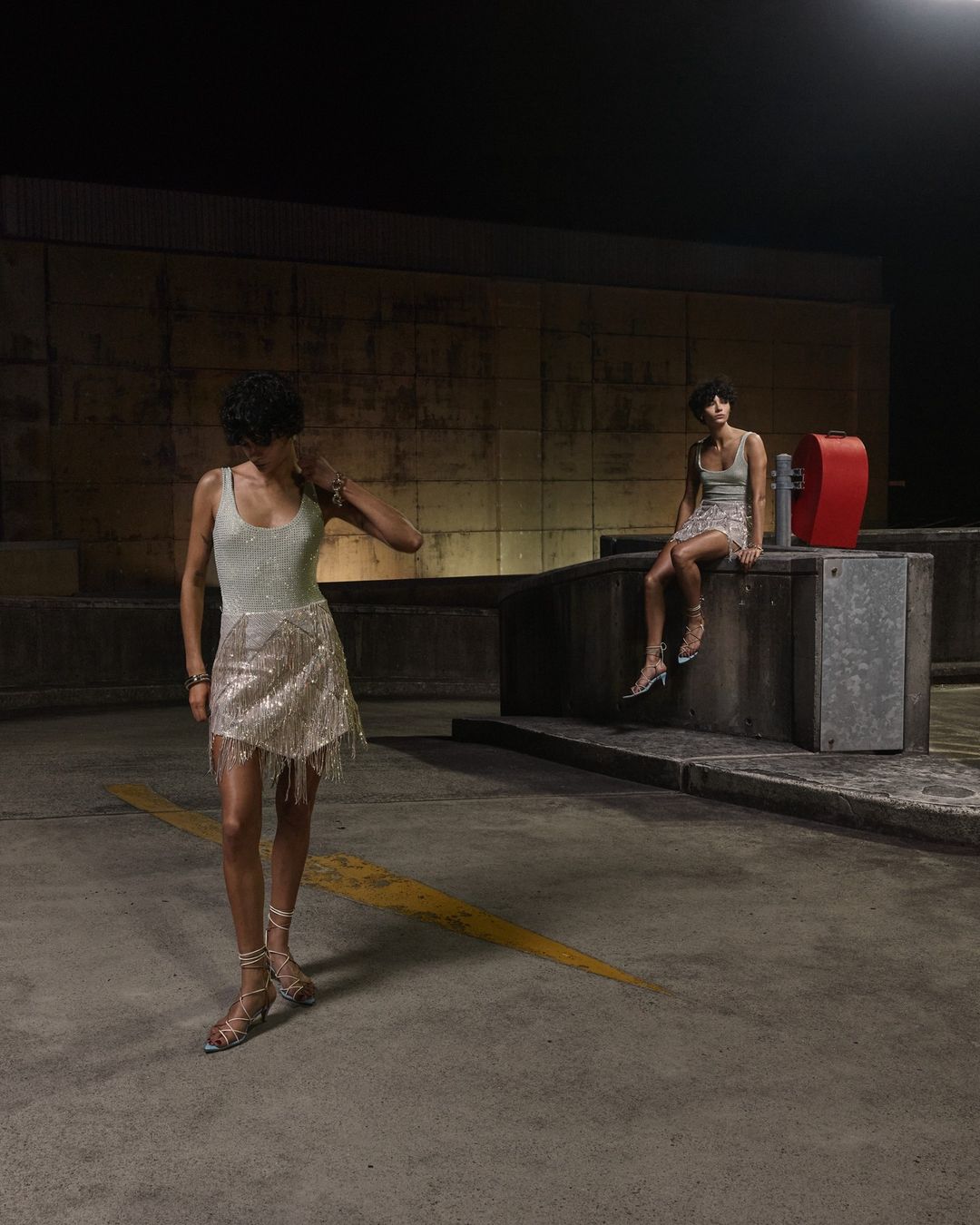 Two young women pose in sequin dresses in an outdoor parking structure with concrete walls and dark lighting, creating an editorial fashion scene.