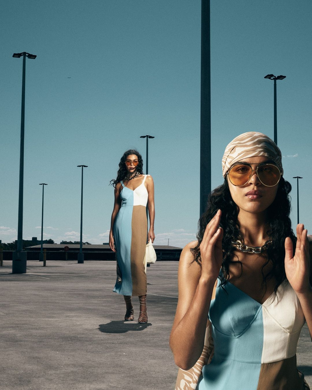 Two women stand on a rooftop parking lot, wearing blue, white, and brown color-block dresses, complemented by tan and gold accessories.