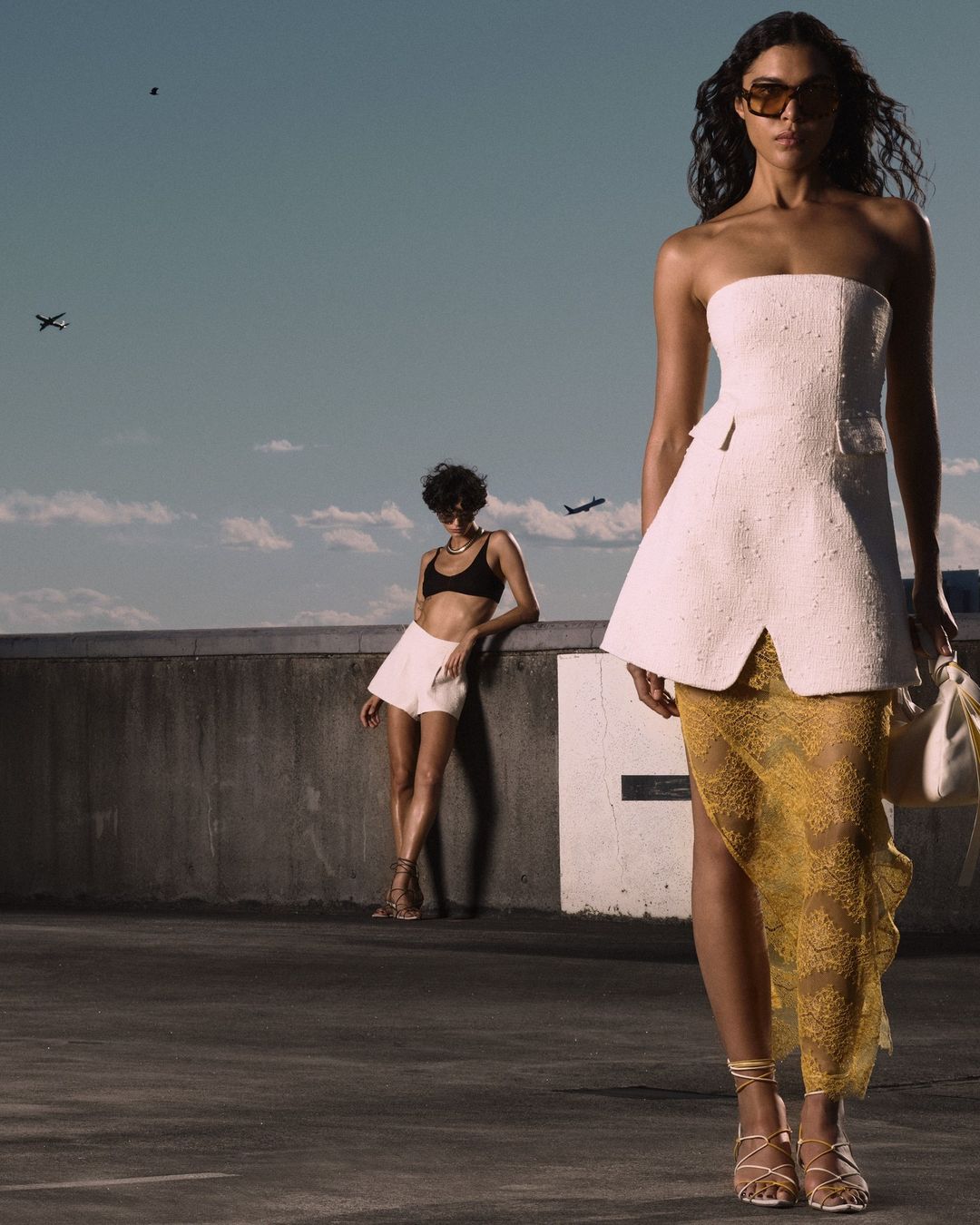 Two fashion models pose on a rooftop wearing strapless tops and dresses, styled with sunglasses, against a blue sky and commercial airplanes.